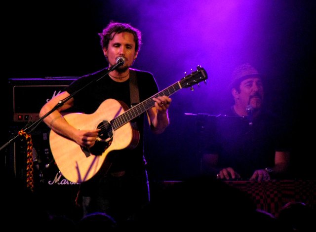 Two musicians on stage under purple light, guitarist at the microphone